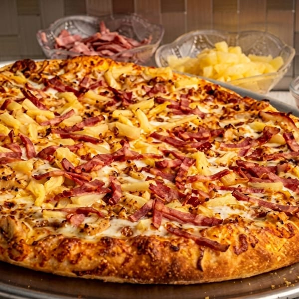 A large Hawaiian pizza with pineapple and ham on a metal tray, with ingredients in bowls.