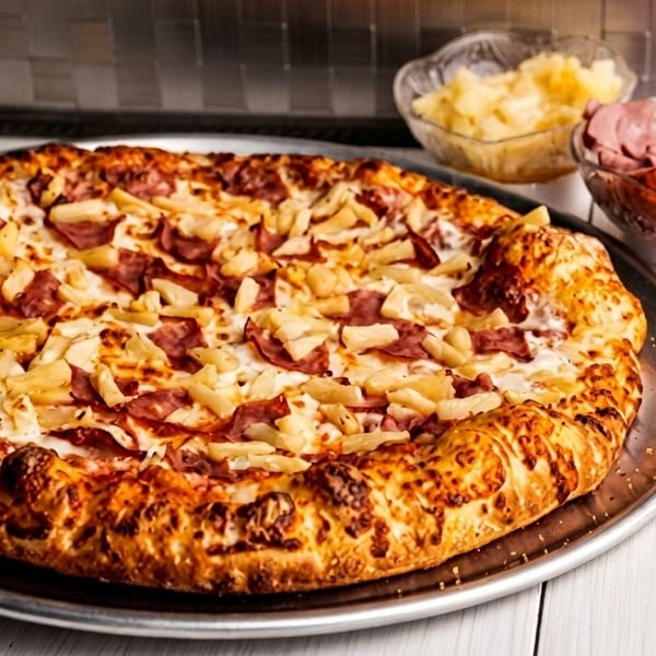 A Hawaiian pizza with ham and pineapple on a metal tray, with bowls of toppings nearby.